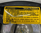 Yellow warning label on a car airbag module against a metallic background